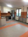 Wnętrze dziennego domu pomocy. Kuchnia. Na podłodze wykładzina typu tarket w kolorach szarym i pomarańczowym. W głębi zabudowa kuchenna ze sprzętem AGD